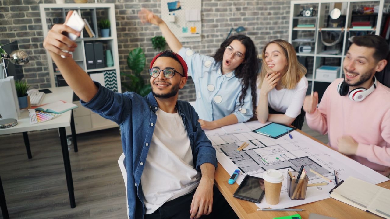 A diverse group of young adults having fun and taking a selfie in a modern office setting.